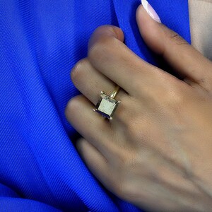 Puede incluir: Un anillo de oro con una piedra cuadrada de color gris oscuro. El anillo est&aacute; en el dedo de una persona.