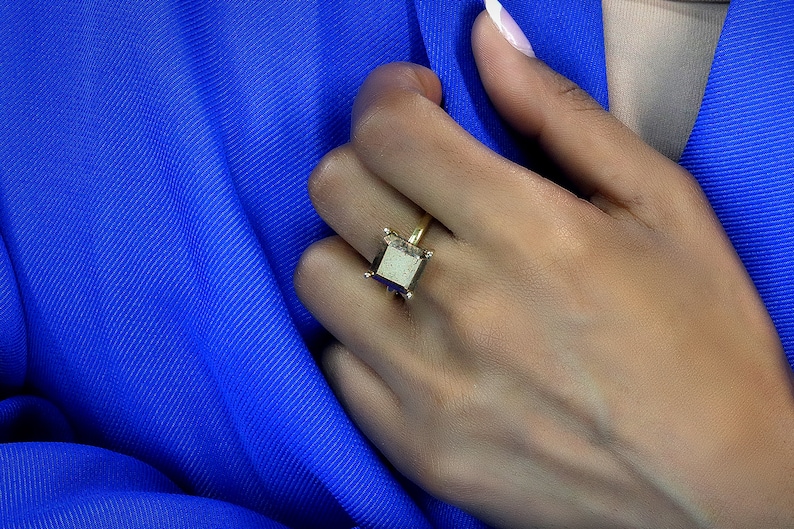 Puede incluir: Un anillo de oro con una piedra cuadrada de color gris oscuro. El anillo est&aacute; en el dedo de una persona.