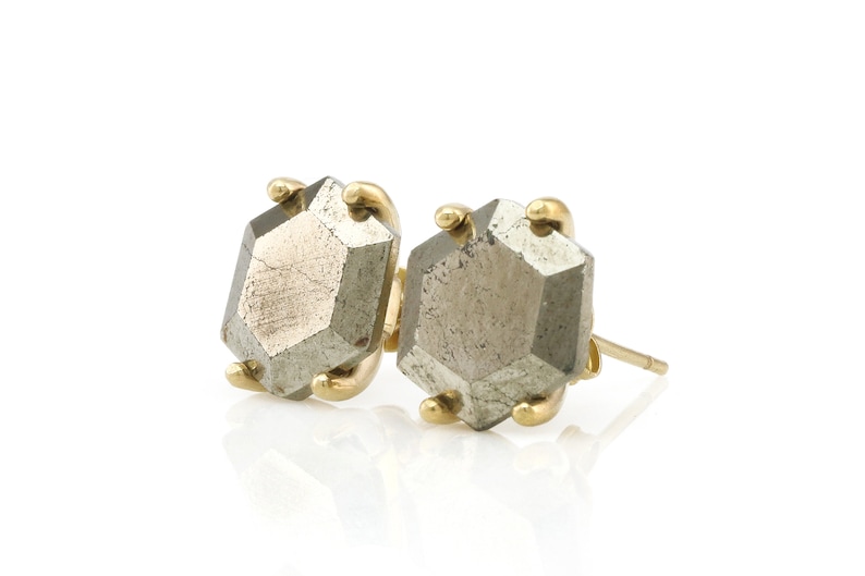 Op de afbeelding: Een paar oorstekers met facetgeslepen, geometrische pyrietstenen. De stenen worden op hun plaats gehouden door goudkleurige klauwen, met gouden pinnen voor doorboorde oren. De oorbellen worden op een witte achtergrond getoond.