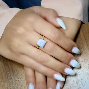 May include: A gold ring with a square-shaped blue agate gemstone. The ring is on a finger with white nail polish.