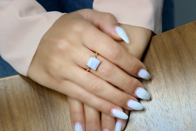 May include: A gold ring with a square-shaped blue agate gemstone. The ring is on a finger with white nail polish.