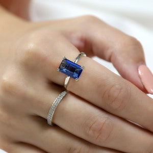 Pode incluir: Um anel de ouro com uma grande gema retangular azul. O anel é usado no dedo indicador de uma mão.