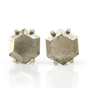 Op de afbeelding: Een paar pyriet oorknopjes. Elke oorbel heeft een gefacetteerde, zeshoekige pyrietsteen in een zilveren zetting. De pyriet heeft een metallic, goudachtige uitstraling. De oorbellen worden op een witte achtergrond gepresenteerd.