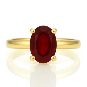 May include: A gold ring featuring a large, oval-cut, deep red gemstone. The gemstone is held in place by four gold prongs. The ring band is a simple, polished gold. The ring is displayed on a white surface with a reflection.