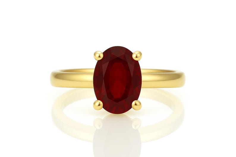 May include: A gold ring featuring a large, oval-cut, deep red gemstone. The gemstone is held in place by four gold prongs. The ring band is a simple, polished gold. The ring is displayed on a white surface with a reflection.