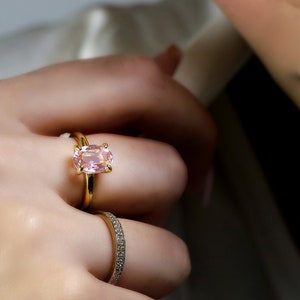 May include: A gold ring with a large oval pink gemstone. The ring is worn on a person's finger, along with a thin gold band with small diamonds.