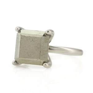Puede incluir: Un anillo de plata con una piedra preciosa de pirita de forma cuadrada. El anillo tiene una banda simple y la piedra preciosa est&aacute; engastada en un engaste de bisel.