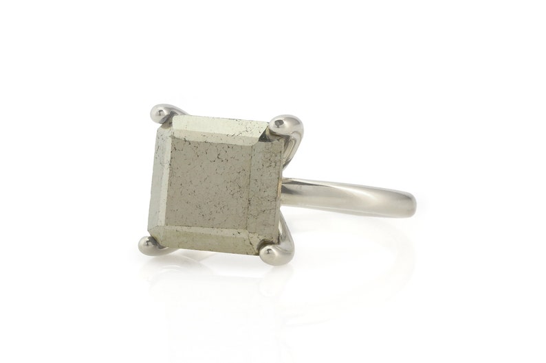 Puede incluir: Un anillo de plata con una piedra preciosa de pirita de forma cuadrada. El anillo tiene una banda simple y la piedra preciosa est&aacute; engastada en un engaste de bisel.