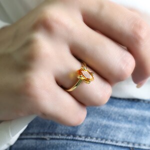 May include: A gold ring featuring an oval-cut, orange gemstone. The ring has a delicate, floral-inspired design around the gemstone. The ring is set on a slender gold band. The ring is shown on a person's finger.