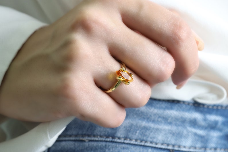May include: A gold ring featuring an oval-cut, orange gemstone. The ring has a delicate, floral-inspired design around the gemstone. The ring is set on a slender gold band. The ring is shown on a person's finger.