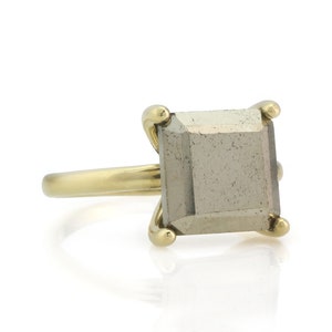 Puede incluir: Un anillo de oro con una piedra preciosa de pirita de forma cuadrada. El anillo tiene una banda simple y la piedra preciosa est&aacute; engastada en un engaste de bisel.