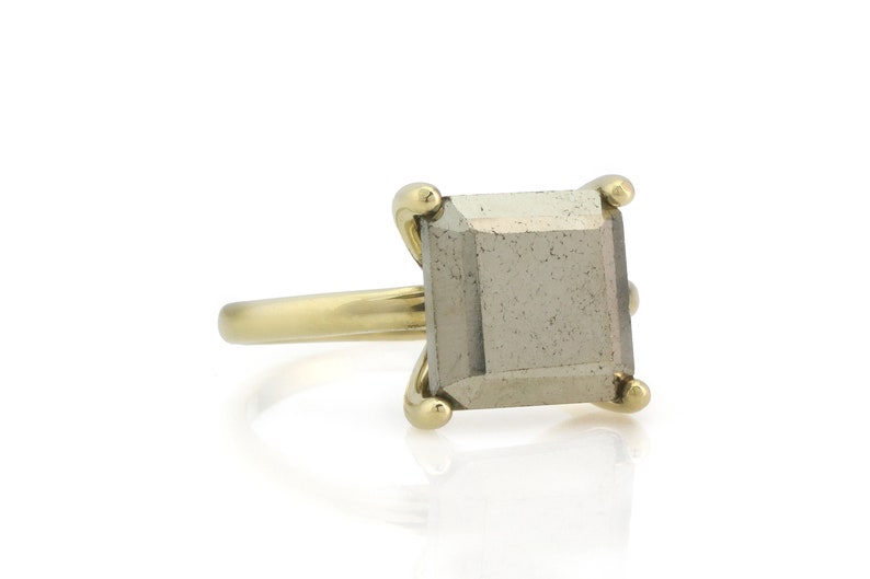 Puede incluir: Un anillo de oro con una piedra preciosa de pirita de forma cuadrada. El anillo tiene una banda simple y la piedra preciosa est&aacute; engastada en un engaste de bisel.