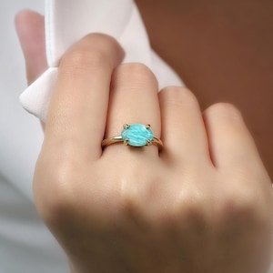 May include: A gold ring featuring a large, light blue gemstone. The ring's band is a simple, thin design, and the gemstone is set with four prongs. The ring is on a person's finger.