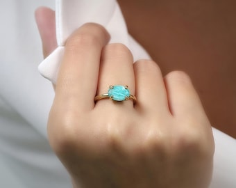 May include: A gold ring featuring a large, light blue gemstone. The ring's band is a simple, thin design, and the gemstone is set with four prongs. The ring is on a person's finger.