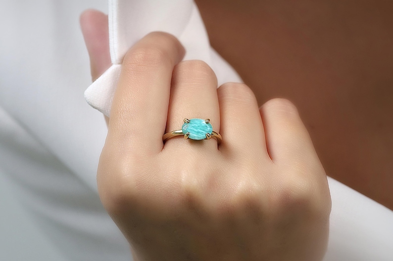 May include: A gold ring featuring a large, light blue gemstone. The ring's band is a simple, thin design, and the gemstone is set with four prongs. The ring is on a person's finger.