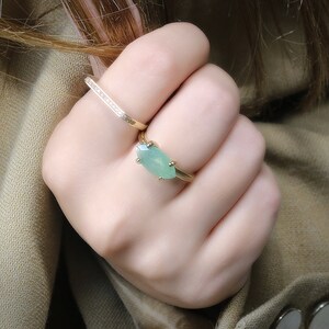 May include: A gold ring with a light green gemstone set in a marquise shape. The ring is worn on a finger with a thin gold band with small diamonds.