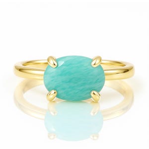 May include: A gold ring featuring a large, oval-shaped, light turquoise gemstone. The gemstone is held in place by four gold prongs. The ring band is a simple, polished gold. The ring is displayed on a white surface with a reflection.