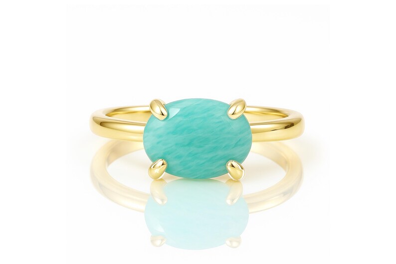 May include: A gold ring featuring a large, oval-shaped, light turquoise gemstone. The gemstone is held in place by four gold prongs. The ring band is a simple, polished gold. The ring is displayed on a white surface with a reflection.