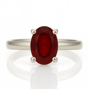 May include: A silver ring featuring a large, oval-cut, deep red gemstone. The gemstone is held in place by four small, round prongs. The ring band is smooth and polished, reflecting light.