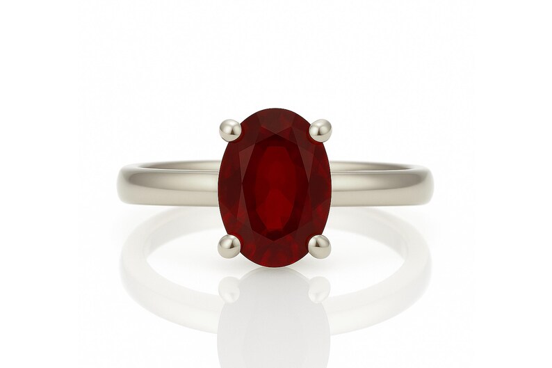 May include: A silver ring featuring a large, oval-cut, deep red gemstone. The gemstone is held in place by four small, round prongs. The ring band is smooth and polished, reflecting light.