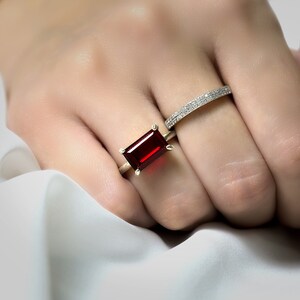 Op de afbeelding: Een gouden ring met een grote, rechthoekige, rode edelsteen. Een dunne, zilveren band met kleine diamanten wordt op dezelfde vinger gedragen.