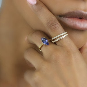 May include: A gold ring featuring a large, blue, marquise-shaped gemstone. The ring is worn on the ring finger of a person's left hand. A second gold ring with a row of small, clear gemstones is worn on the same finger.