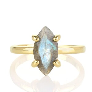 Puede incluir: Un anillo de oro con una piedra de labradorita en forma de marquesa. La piedra preciosa tiene un color base gris con destellos azules y verdes. La banda y las garras del anillo son de color dorado, creando un dise&ntilde;o cl&aacute;sico y elegante.