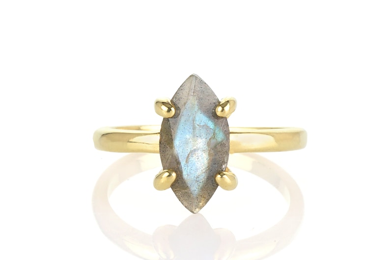 Puede incluir: Un anillo de oro con una piedra de labradorita en forma de marquesa. La piedra preciosa tiene un color base gris con destellos azules y verdes. La banda y las garras del anillo son de color dorado, creando un dise&ntilde;o cl&aacute;sico y elegante.