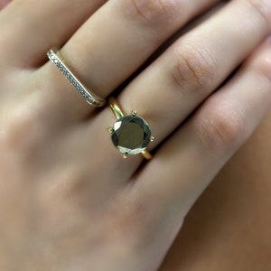 May include: A gold ring with a large, round, dark green gemstone set in a prong setting. The ring is worn on a finger with a thin, silver band with small, clear stones.