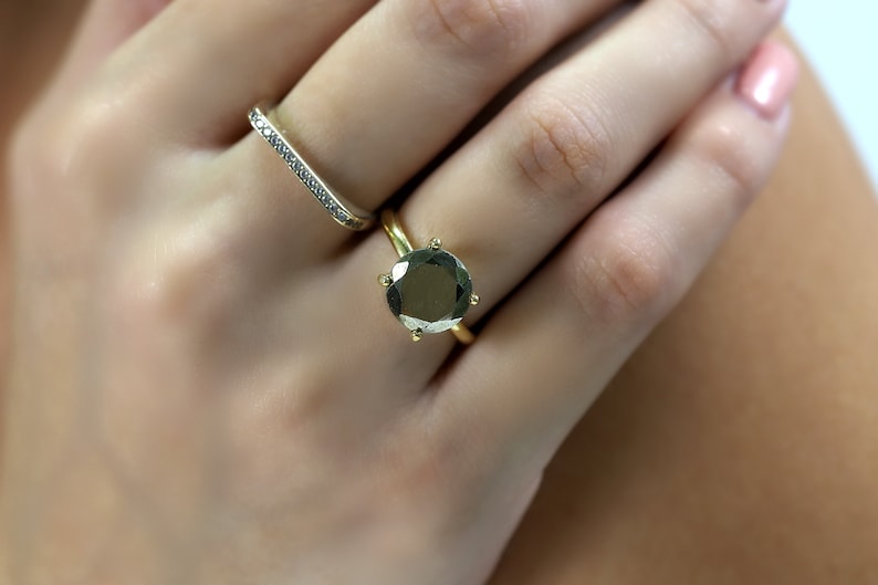 May include: A gold ring with a large, round, dark green gemstone set in a prong setting. The ring is worn on a finger with a thin, silver band with small, clear stones.