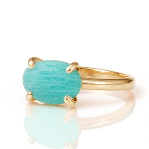 May include: A gold ring featuring a large, oval-shaped, light blue gemstone. The gemstone is held in place by four gold prongs. The ring band is a simple, polished gold. The ring is set against a white background.