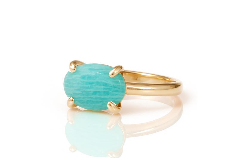 May include: A gold ring featuring a large, oval-shaped, light blue gemstone. The gemstone is held in place by four gold prongs. The ring band is a simple, polished gold. The ring is set against a white background.