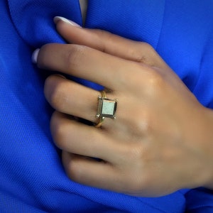 Puede incluir: Un anillo de oro con una piedra preciosa de forma cuadrada. La piedra preciosa es de color gris claro con un borde gris oscuro.