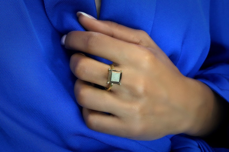 Puede incluir: Un anillo de oro con una piedra preciosa de forma cuadrada. La piedra preciosa es de color gris claro con un borde gris oscuro.