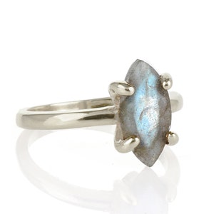 Puede incluir: Un anillo de plata con una piedra de labradorita en forma de marquesa. La piedra gris tiene destellos azules e iridiscentes. El anillo tiene una banda sencilla y cuatro garras que sujetan la piedra.