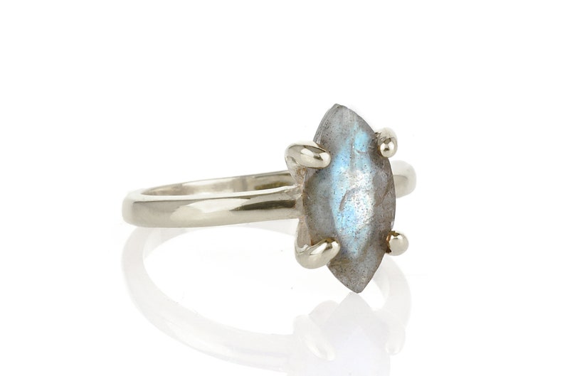 Puede incluir: Un anillo de plata con una piedra de labradorita en forma de marquesa. La piedra gris tiene destellos azules e iridiscentes. El anillo tiene una banda sencilla y cuatro garras que sujetan la piedra.