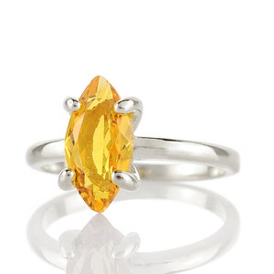 May include: A silver ring featuring a marquise-cut, bright yellow gemstone. The gemstone is held in place by four silver prongs. The ring is set against a white background, with a reflection visible below.