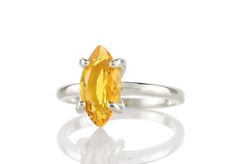 May include: A silver ring featuring a marquise-cut, bright yellow gemstone. The gemstone is held in place by four silver prongs. The ring is set against a white background, with a reflection visible below.