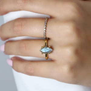 Puede incluir: Se muestra un anillo de oro con una piedra preciosa iridiscente de talla marquesa. Tambi&eacute;n es visible un segundo anillo con peque&ntilde;as piedras transparentes. Los anillos est&aacute;n en el dedo de una persona, con un fondo suave.