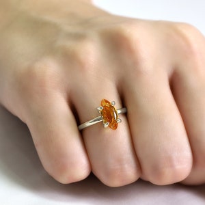 May include: A silver ring with a marquise-cut, orange gemstone. The gemstone is set in a gold bezel and has a pointed shape. The ring is displayed on a person's hand.