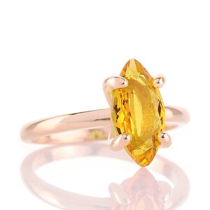 May include: A rose gold ring featuring a marquise-cut, bright yellow gemstone. The gemstone is held in place by four prongs. The ring is set against a white background, with a reflection visible below.