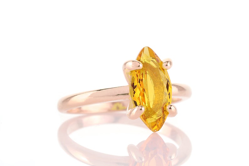 May include: A rose gold ring featuring a marquise-cut, bright yellow gemstone. The gemstone is held in place by four prongs. The ring is set against a white background, with a reflection visible below.