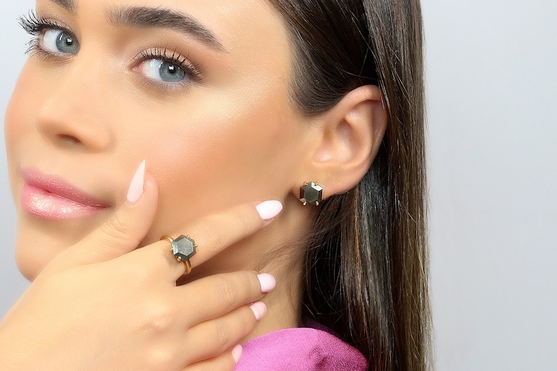 Op de afbeelding: Een gouden ring en bijpassende oorstekers met zeshoekige, donkergrijze edelstenen. De ring zit om een vinger en de oorbellen zitten in een oor. De sieraden staan tegen een lichte achtergrond.