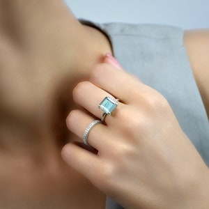 Könnte beinhalten: Ein silberner Ring mit einem großen, quadratischen, hellblauen Edelstein. Der Ring wird am Ringfinger einer Hand getragen.