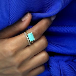 May include: A hand wearing two gold rings. One ring features a rectangular, light blue gemstone. The other ring is a thin band with small, clear stones. The background is a vibrant blue fabric.