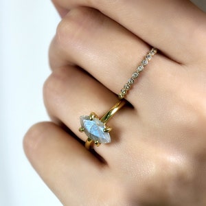 Puede incluir: Un anillo de oro con una piedra preciosa en forma de marquesa, de color azul claro y blanco. Tambi&eacute;n se ve un segundo anillo de oro con peque&ntilde;as piedras transparentes. Los anillos est&aacute;n en una mano.