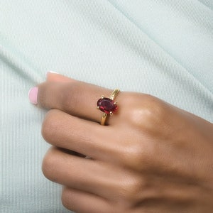 May include: A gold ring featuring a large, oval-shaped, deep red gemstone. The ring is set on a slender gold band and is displayed on a hand against a light green background. The gemstone is the focal point, catching the light.