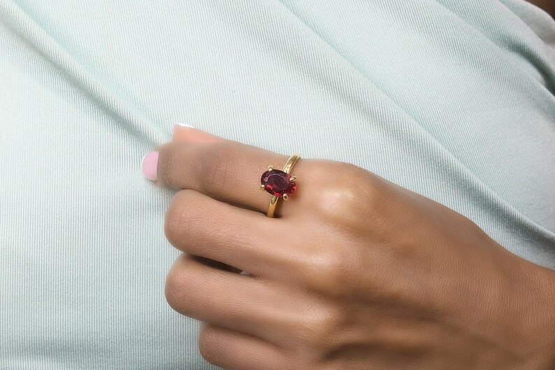 May include: A gold ring featuring a large, oval-shaped, deep red gemstone. The ring is set on a slender gold band and is displayed on a hand against a light green background. The gemstone is the focal point, catching the light.