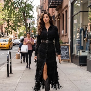 May include: A long-sleeved black suede shirt dress featuring multiple horizontal tiers of fringe detailing, a button-down front closure, a classic collar, and a black leather belt with a silver-toned buckle, paired with dark leather cowboy boots.