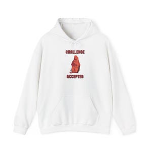 May include: A white long-sleeve hooded sweatshirt featuring a central graphic of a red hooded figure. The text "CHALLENGE" is printed in red above the figure, and the word "ACCEPTED" is printed in red below the figure. The hoodie has a front pocket.